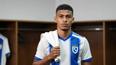 A 26-year-old male midfielder in a white and blue jersey, with a determined expression, standing in a locker room with a...