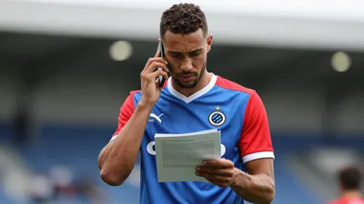 Mid-30s athletic forward, waist-up, wearing Club Brugge home jersey - vivid blue body with red sleeves, crisp white...