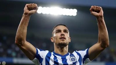 A Club Brugge player in a blue and white striped shirt celebrating a goal, arms raised