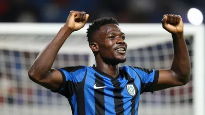 A tight action shot of a young, athletic African midfielder in a blue and black striped jersey, celebrating a goal with...