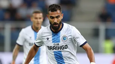 A 25-year-old, athletic, dark-haired, bearded, Club Brugge midfielder in a white jersey with a blue stripe, playing a...